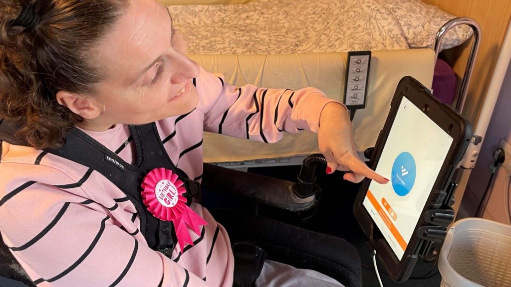A middle aged woman is using the Voiceitt app on an iPad mounted onto her wheelchair